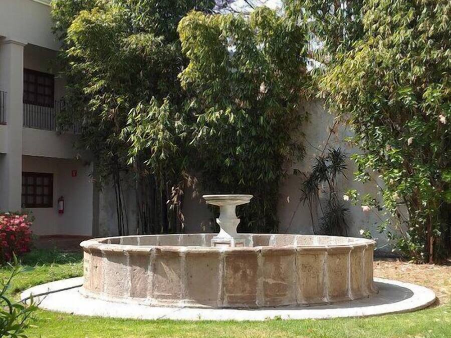 A circular stone fountain with a central pedestal, surrounded by a grassy area with trees and a building in the background.