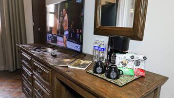 A wooden desk with a television, remote controls, notebooks, water bottles, coffee maker, and a mirror.