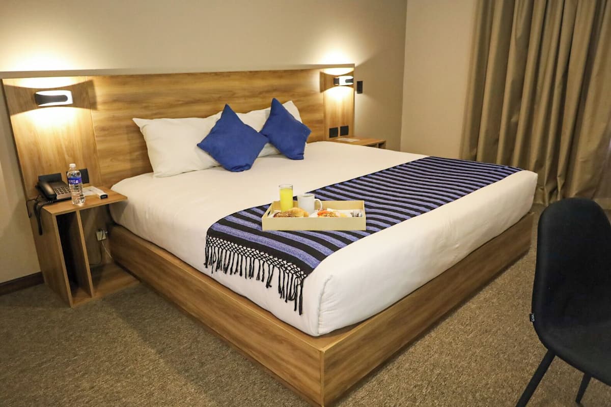 Habitación de hotel con cabecera de madera, dos almohadas azules, una colcha a rayas, una mesita de noche con una botella y un teléfono, y una bandeja con alimentos para el desayuno.