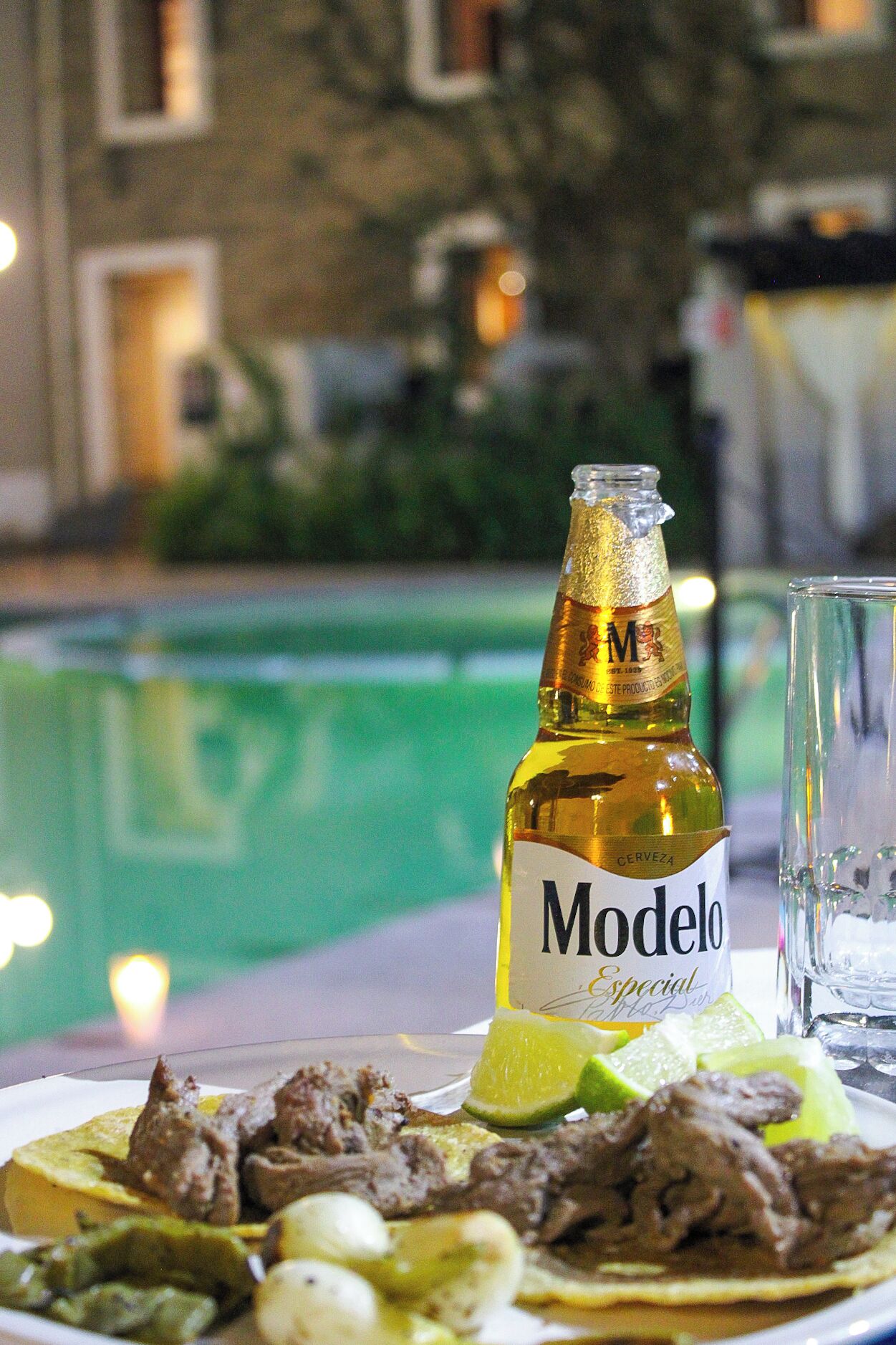 A bottle of Modelo beer, a glass, and a plate with food and lime on a table by a pool.
