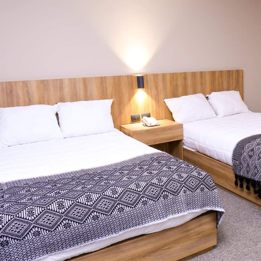A hotel room with two beds, a wooden headboard, a nightstand with a lamp, and a patterned throw blanket.