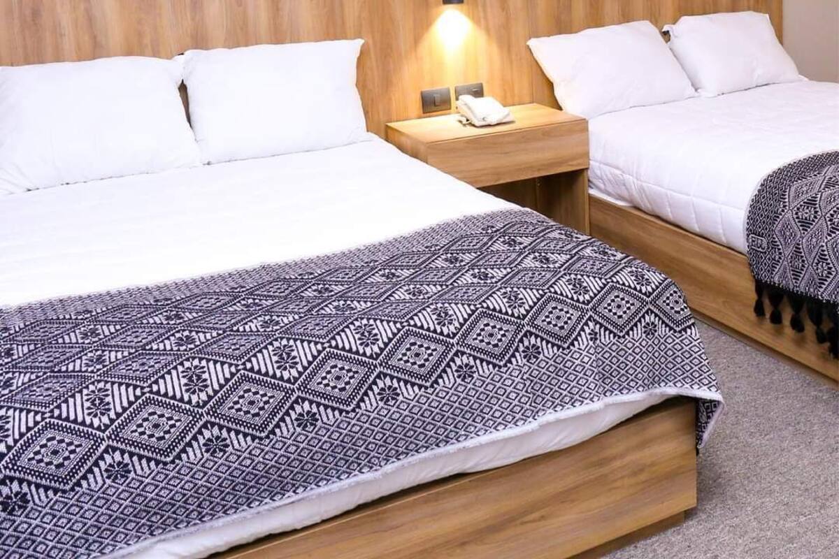 A hotel room with two beds, a wooden headboard, a nightstand with a lamp, and a patterned throw blanket.