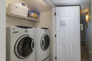 Una sala de lavandería con dos lavadoras de carga frontal, un estante con cestos de ropa y una puerta con un cartel.