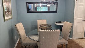 A dining area with a round glass table, four upholstered chairs with metal studs, a chandelier, a framed picture on the wall, and a white door.