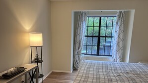 A bedroom with a bed, a bedside table with a lamp, a window with curtains, and a view of greenery outside.