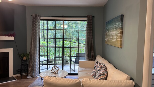 A living room with a beige sofa, a small round table, and a view of greenery through a large window with curtains.