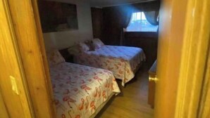 Passadís estret que condueix a una habitació amb dos llits, terres de fusta i una finestra amb cortines.