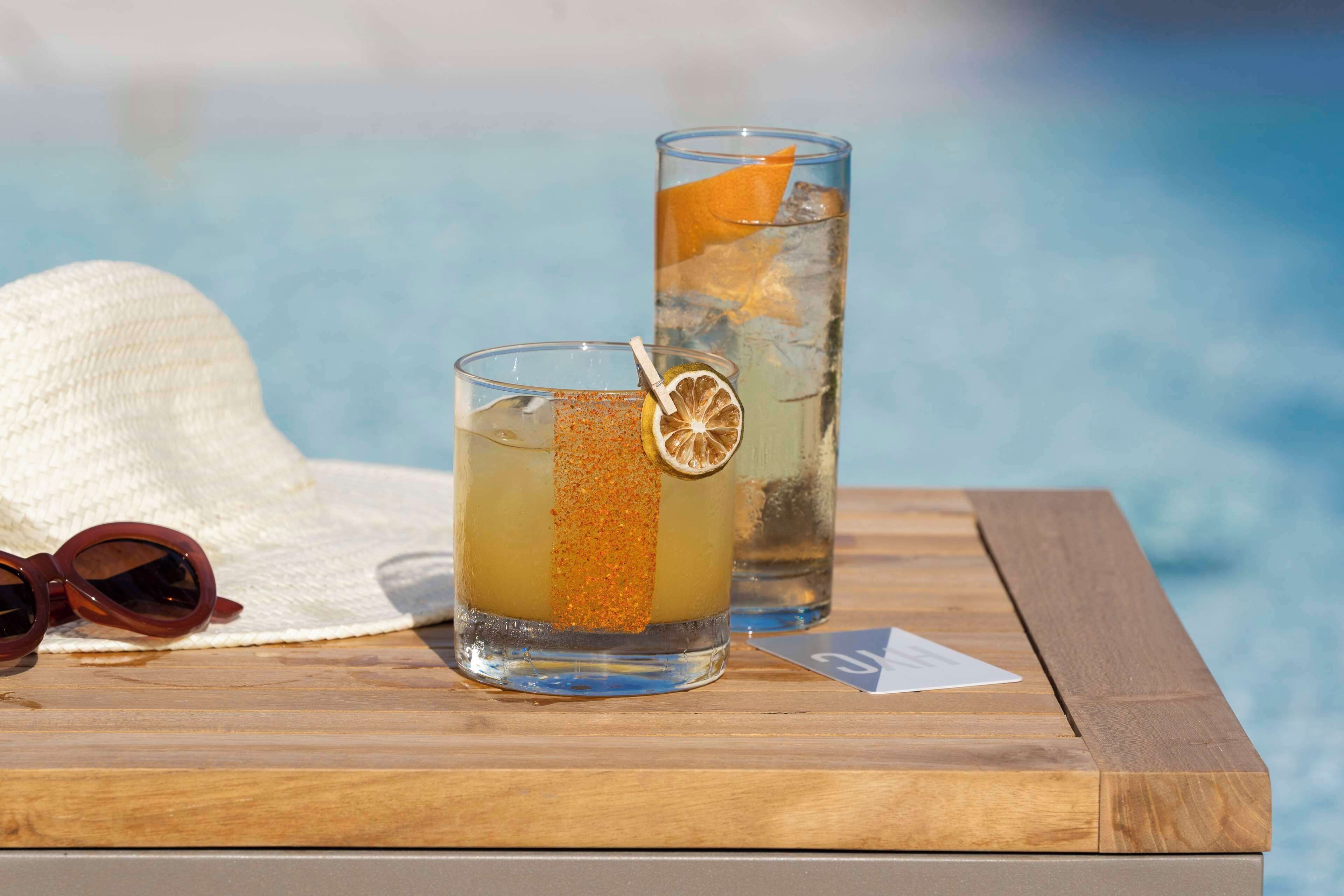 Two glasses of a beverage with a slice of orange and a straw, placed on a wooden table with a hat and sunglasses beside them.