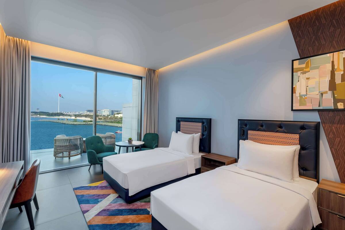 A hotel room with two beds, a desk, a chair, a small table, and a view of the sea.