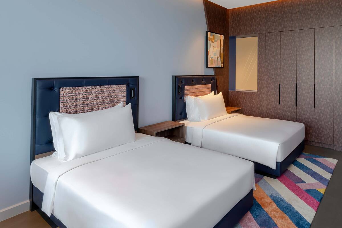 A hotel room with two beds, a wooden nightstand, a colorful rug, and a wardrobe with a geometric-patterned wall.