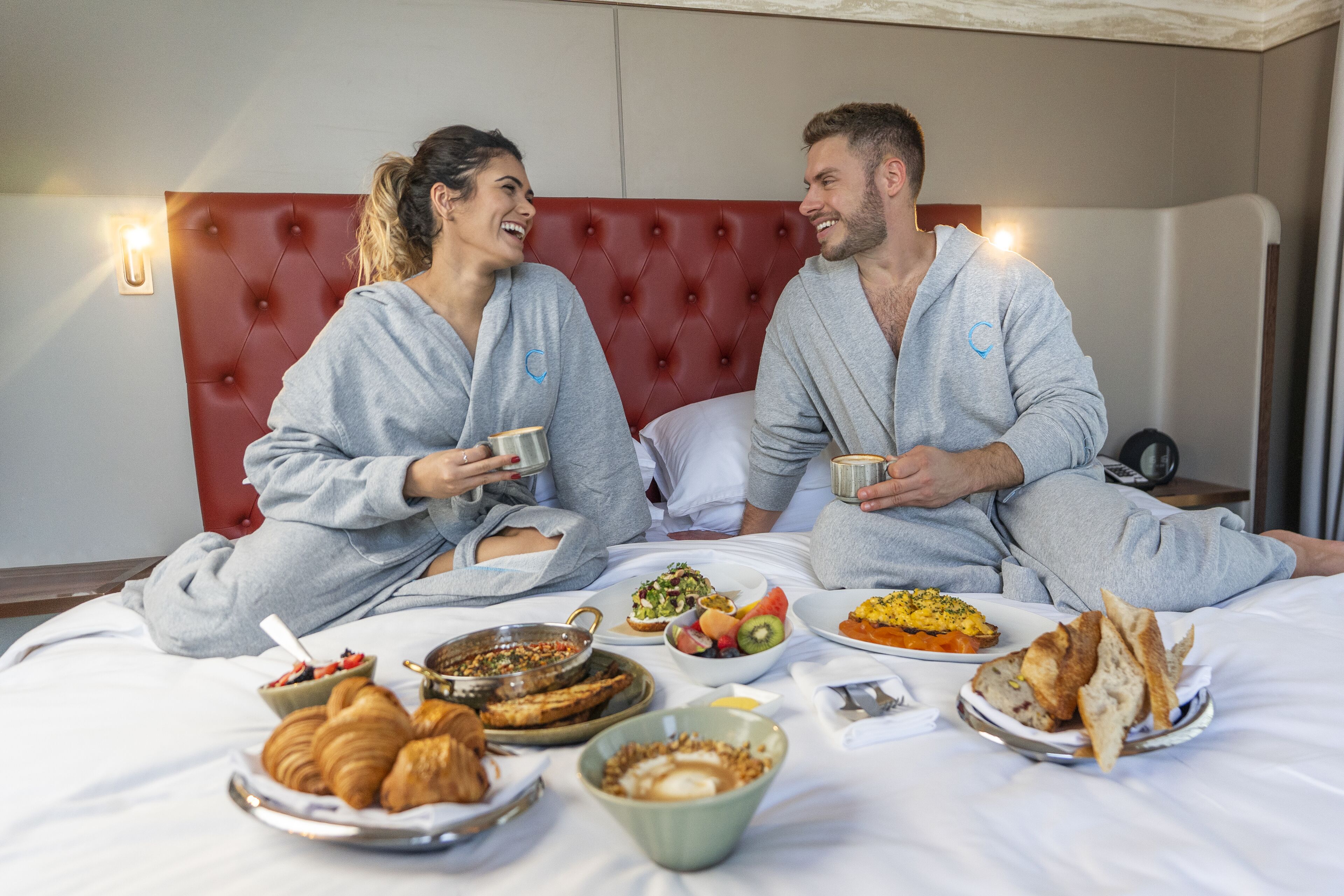A couple sitting on a bed with breakfast items, including croissants, fruit, and eggs.
