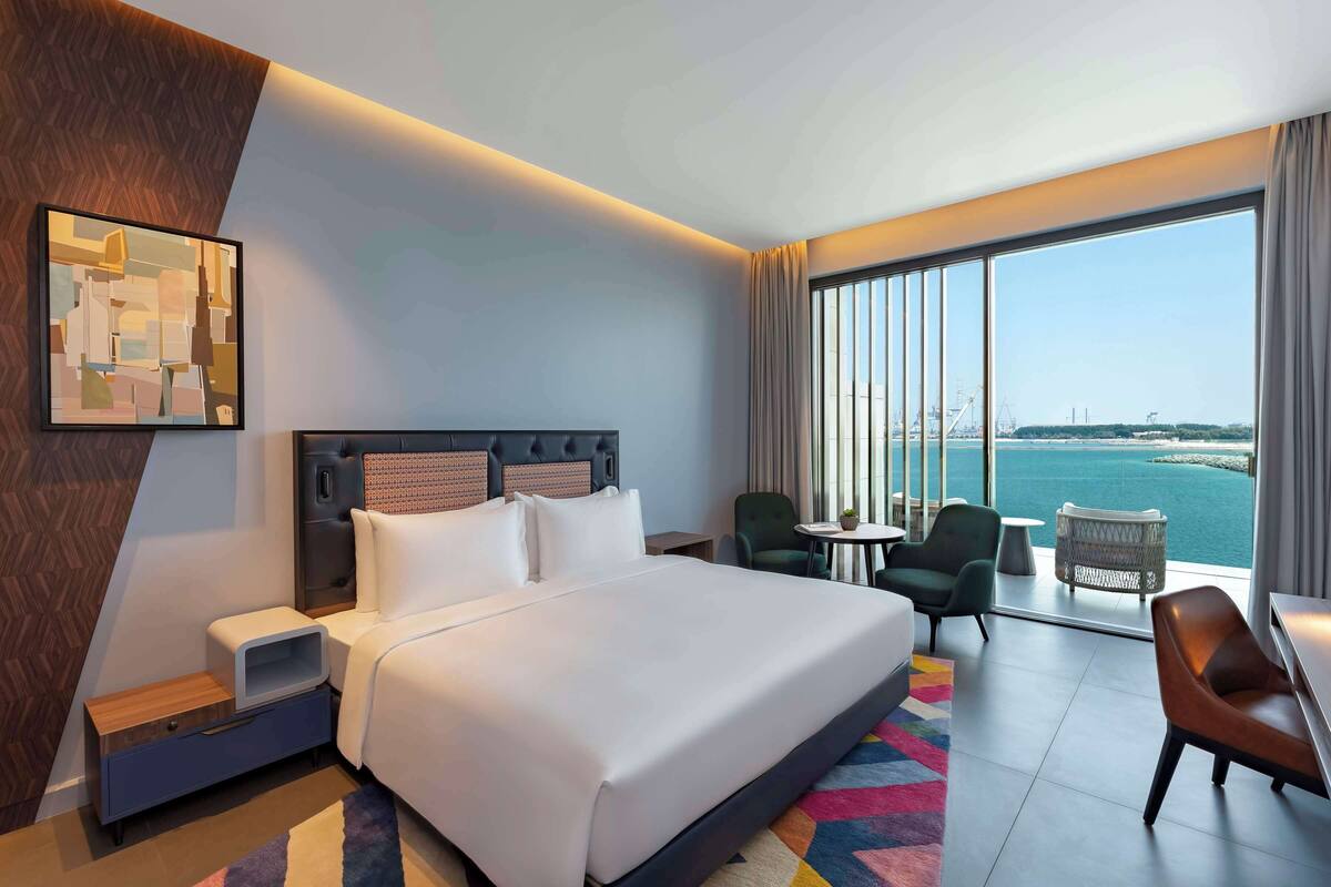 A hotel room with a large bed, a nightstand, a chair, a small table, and a view of the sea.