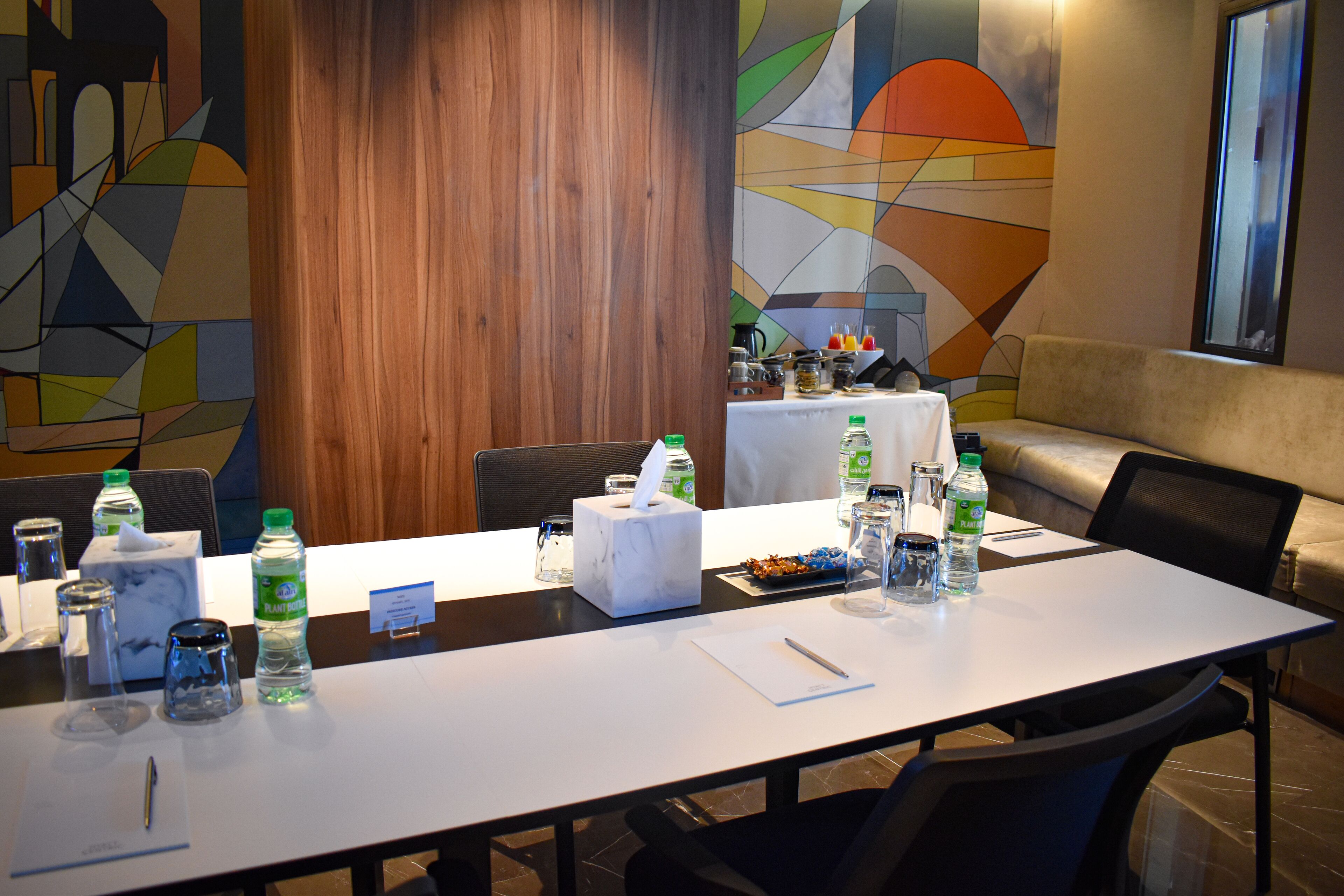 Un salón de conferencias con una larga mesa dispuesta para reuniones, equipado con botellas de agua, una caja de pañuelos y un bloc de notas.