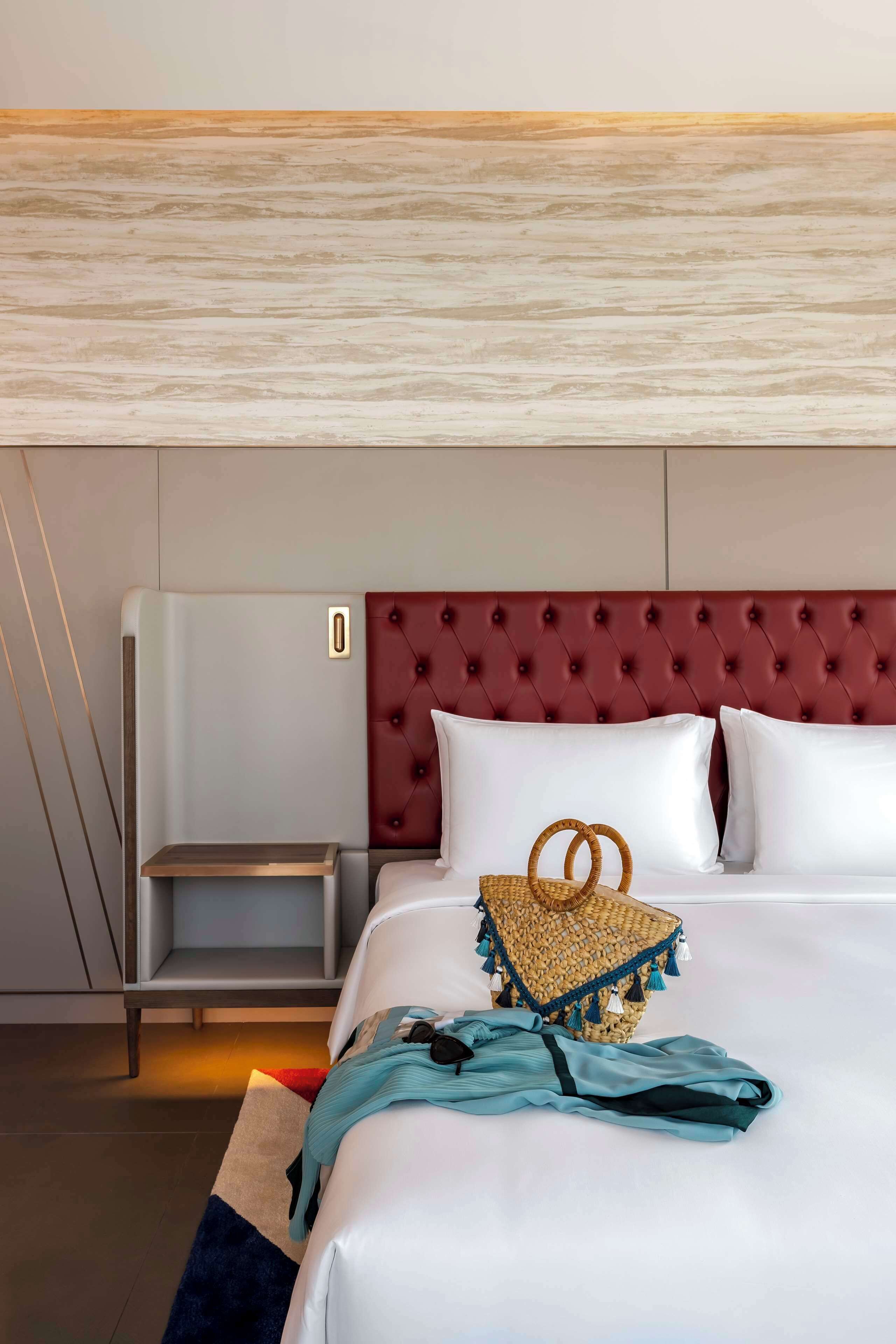 A hotel room with a large bed, a red tufted headboard, a bedside table, and a small wicker basket.