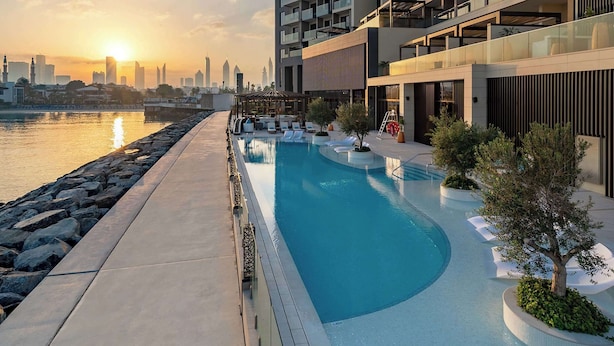 Un hotel moderno con piscina y un horizonte urbano al fondo, bajo un atardecer.