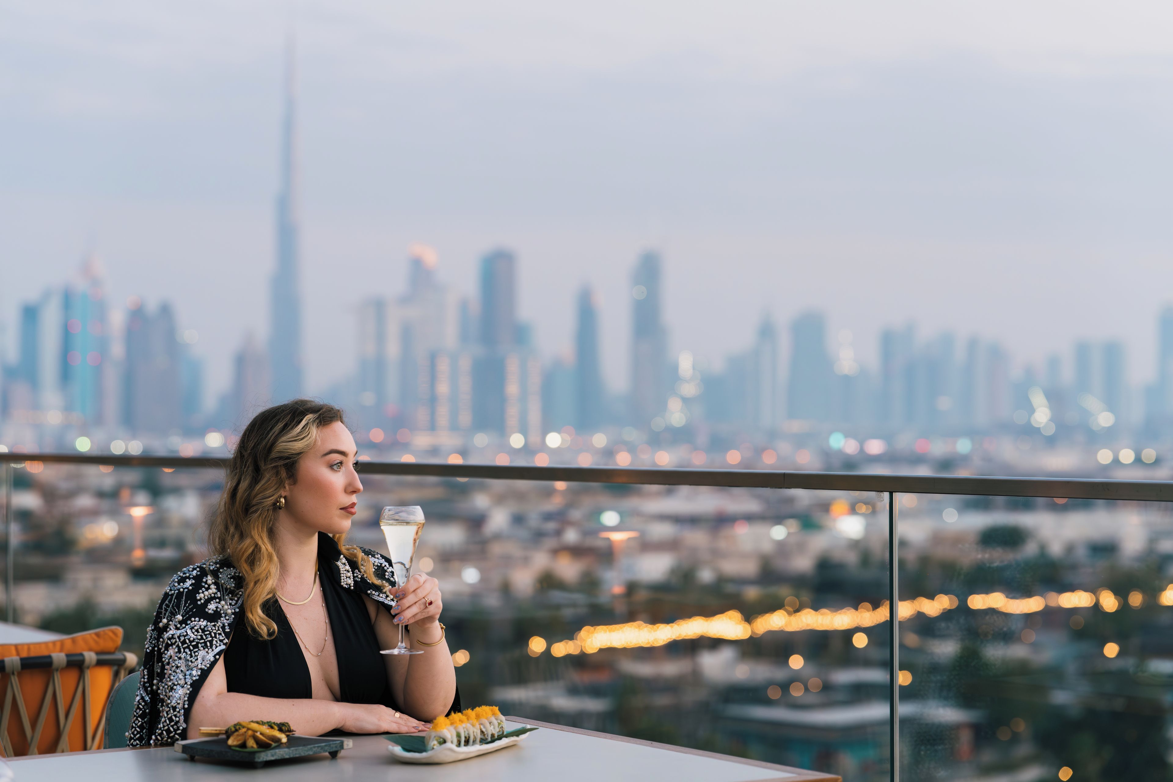 Une personne assise à une table, avec une vue sur la ville, tenant un verre à la main.