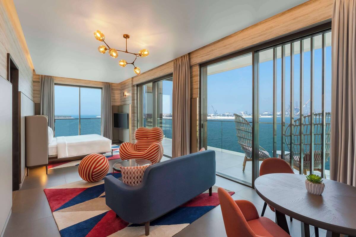A modern hotel room with a large balcony, a sofa, a dining table, and a TV.