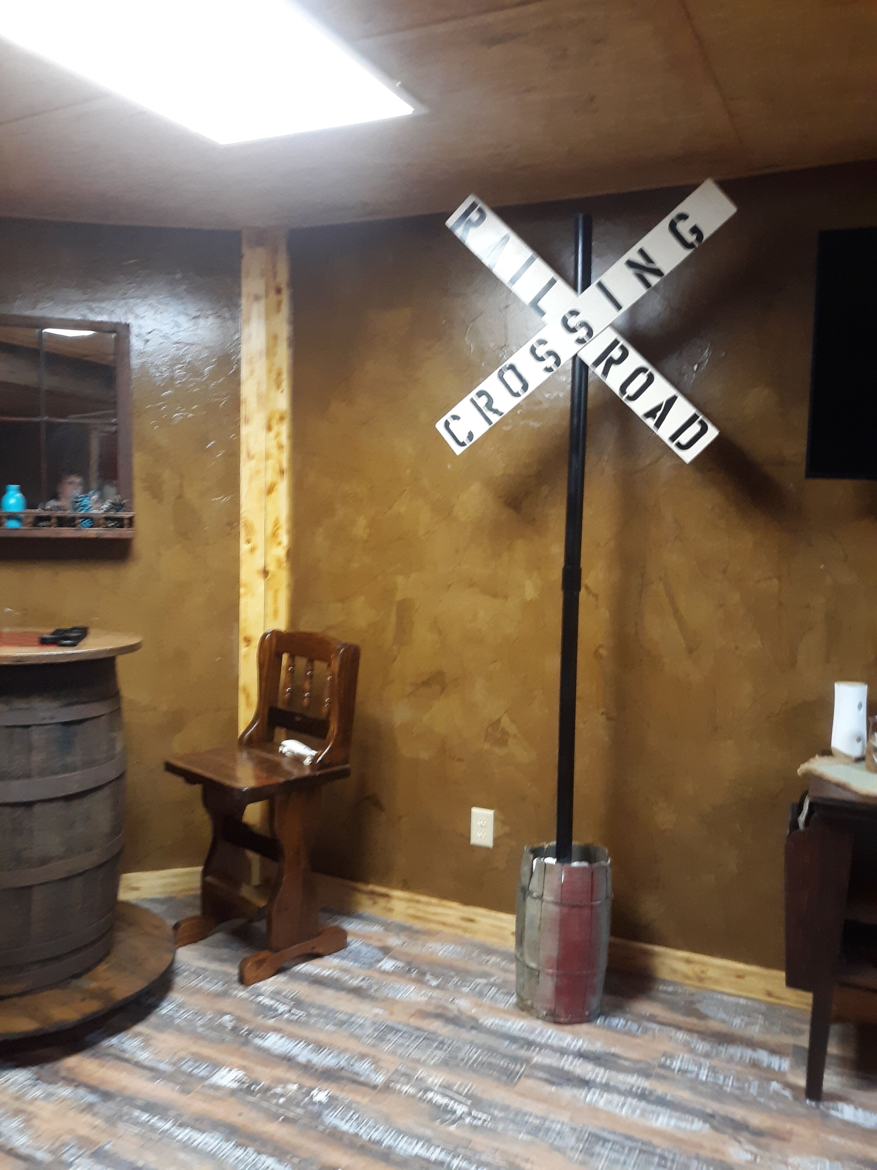 Une pièce avec un mur en bois, une chaise en bois, une petite table et un tonneau. Il y a une pancarte sur le mur où l’on peut lire : « PASSAGE PIÉTON ».