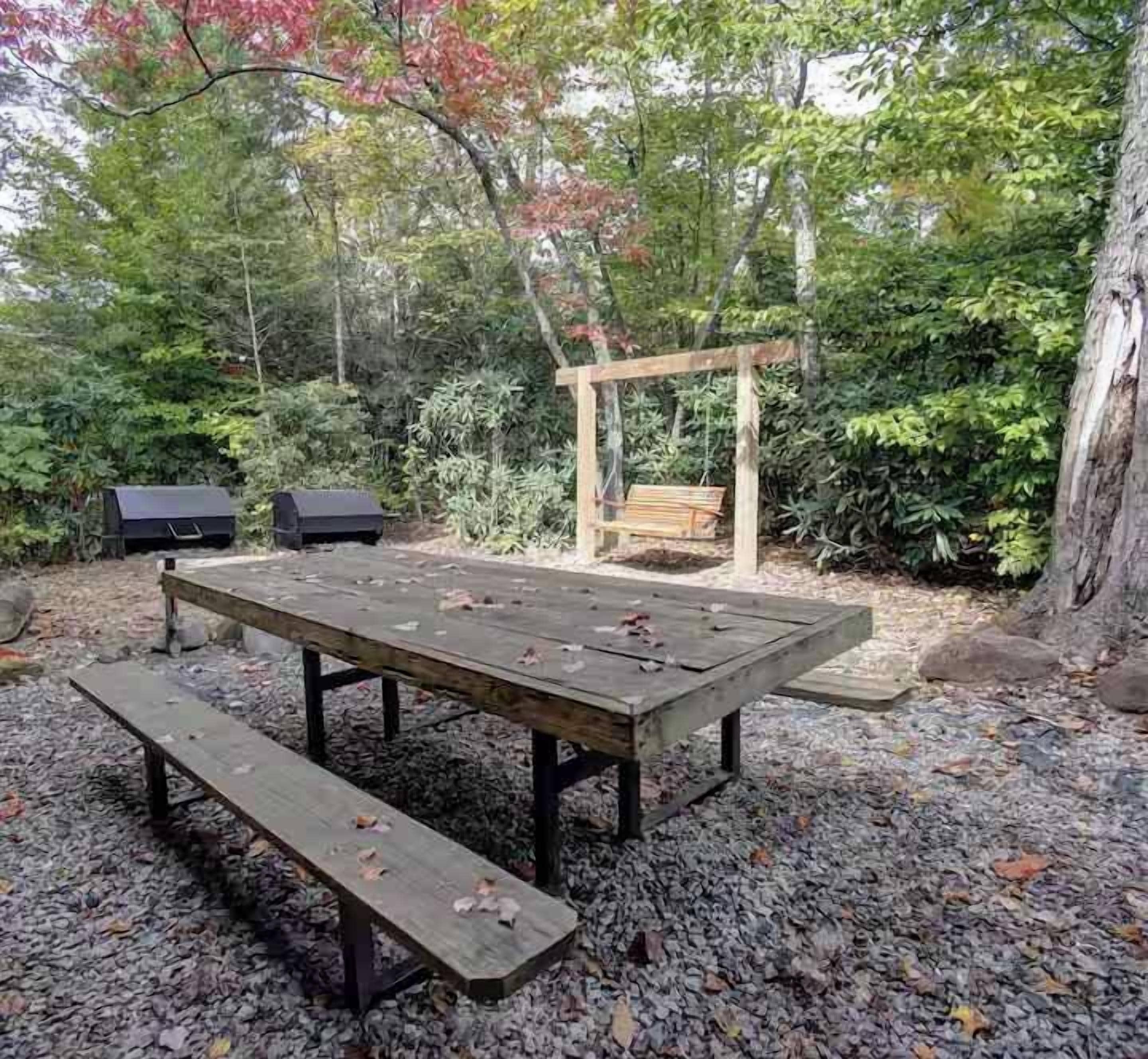 An outdoor seating area with a wooden table and benches, surrounded by trees and foliage.