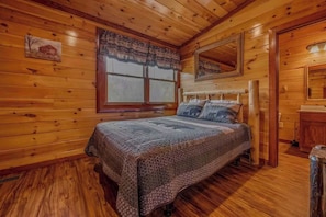 A wooden cabin bedroom with a bed, a window with curtains, a mirror, and a wooden floor. - Grand Bear (Cosby)