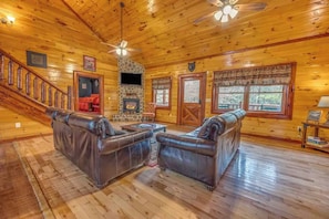 A log cabin interior with a fireplace, leather sofas, a staircase, and a ceiling fan. - Grand Bear (Cosby)
