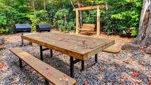 Een buitenruimte met een houten tafel en bankjes, omgeven door grind en herfstbladeren.