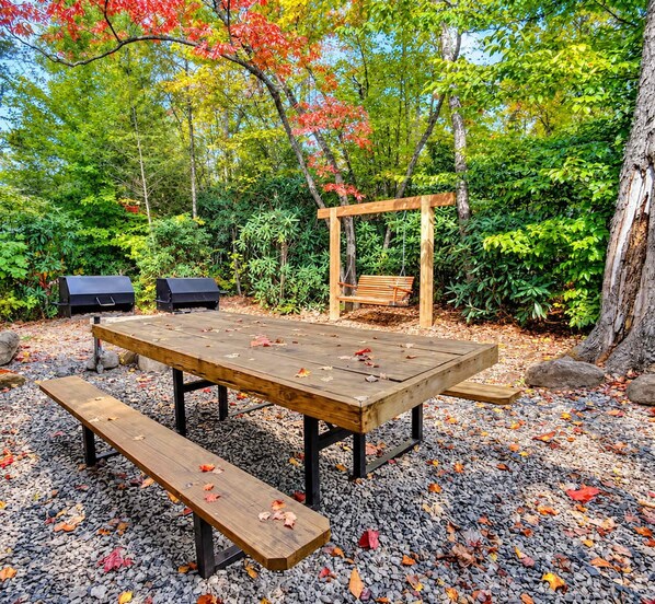 An outdoor seating area with a wooden table and benches, surrounded by gravel and autumn leaves.