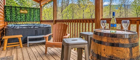 A wooden deck with a hot tub, a wooden chair, a stool, and a barrel with glasses and a bottle.