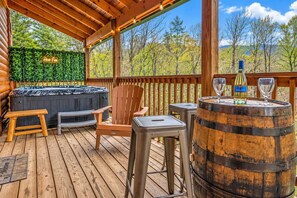 A wooden deck with a hot tub, a wooden chair, a stool, and a barrel with glasses and a bottle.