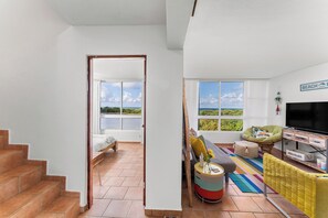 A spacious living area with a large window offering a view of the sea, a comfortable sofa, a television, and a staircase leading to another room. - Walk-up penthouse w/breathtaking views, WiFi, A/C, free laundry, sleeps 6 (Cabo Rojo)