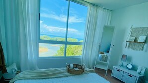 A bedroom with a large bed, a window with a view of the outdoors, a bedside table with a lamp, and a wall-mounted shelf. - Walk-up penthouse w/breathtaking views, WiFi, A/C, free laundry, sleeps 6 (Cabo Rojo)