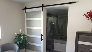 A room with a sliding glass door leading to a bathroom, a grey armchair, a potted plant, and a wooden dresser. - 3 BR Palm Springs Condo. Pool, Hot Tub. Palm Springs Art District. (Palm Springs)
