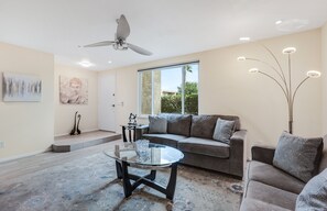 A modern living room with a sofa, a glass coffee table, a ceiling fan, and a large window.