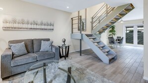 A modern living room with a grey sofa, a glass coffee table, and a staircase leading to an upper level. - 3 BR Palm Springs Condo. Pool, Hot Tub. Palm Springs Art District. (Palm Springs)