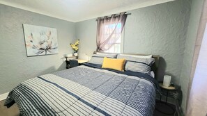 A bedroom with a bed, a bedside table, a nightstand, a window with curtains, a wall-mounted painting, and a vase with flowers.