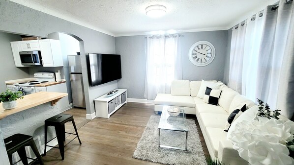 A modern kitchen with a flat-screen TV, a dining area with a wooden table, and a living room with a white sofa and a decorative plant.