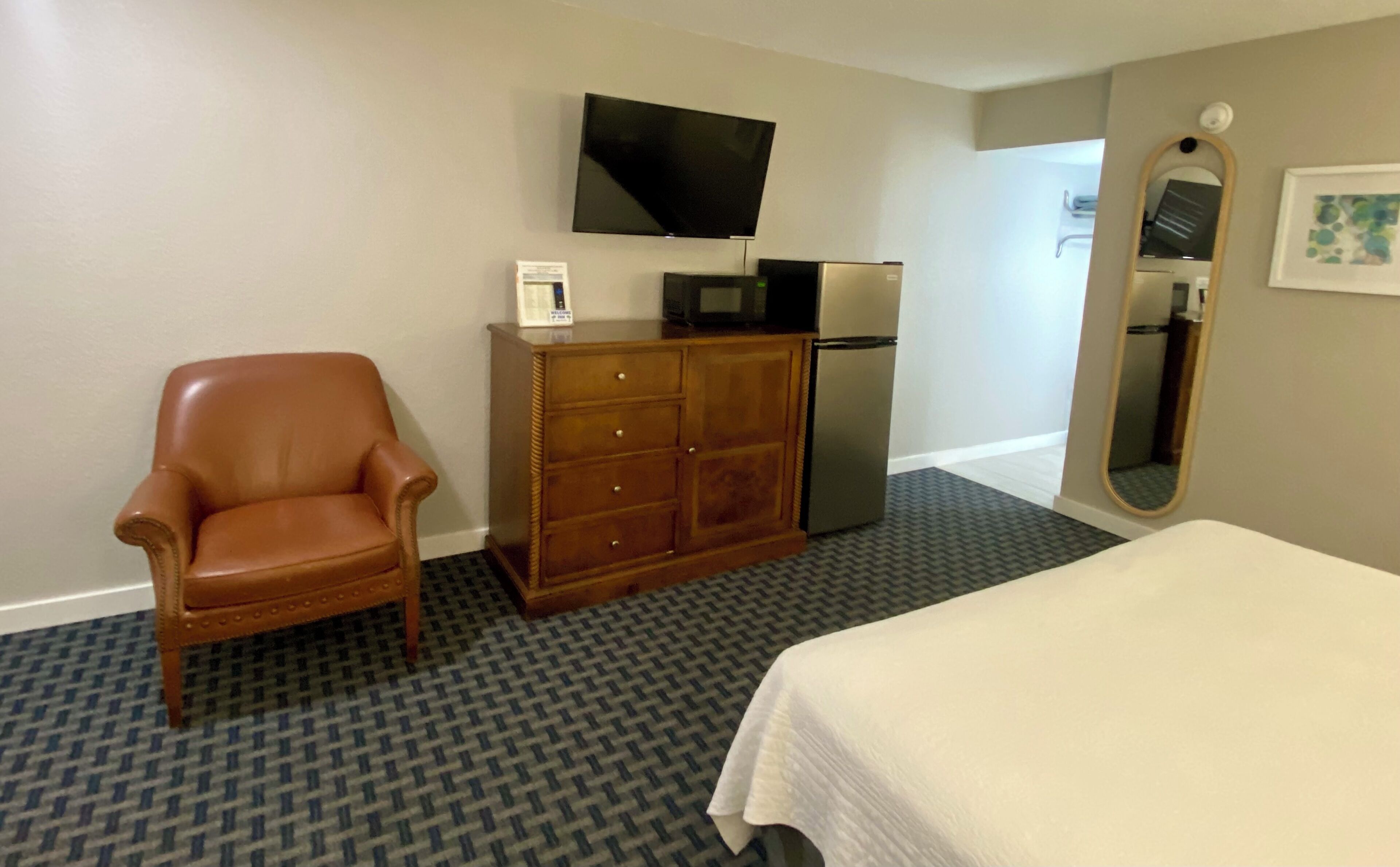 A hotel room with a brown leather chair, a wooden dresser with a flat-screen TV mounted above it, a refrigerator, a bed with a white bedspread, and a patterned carpet.