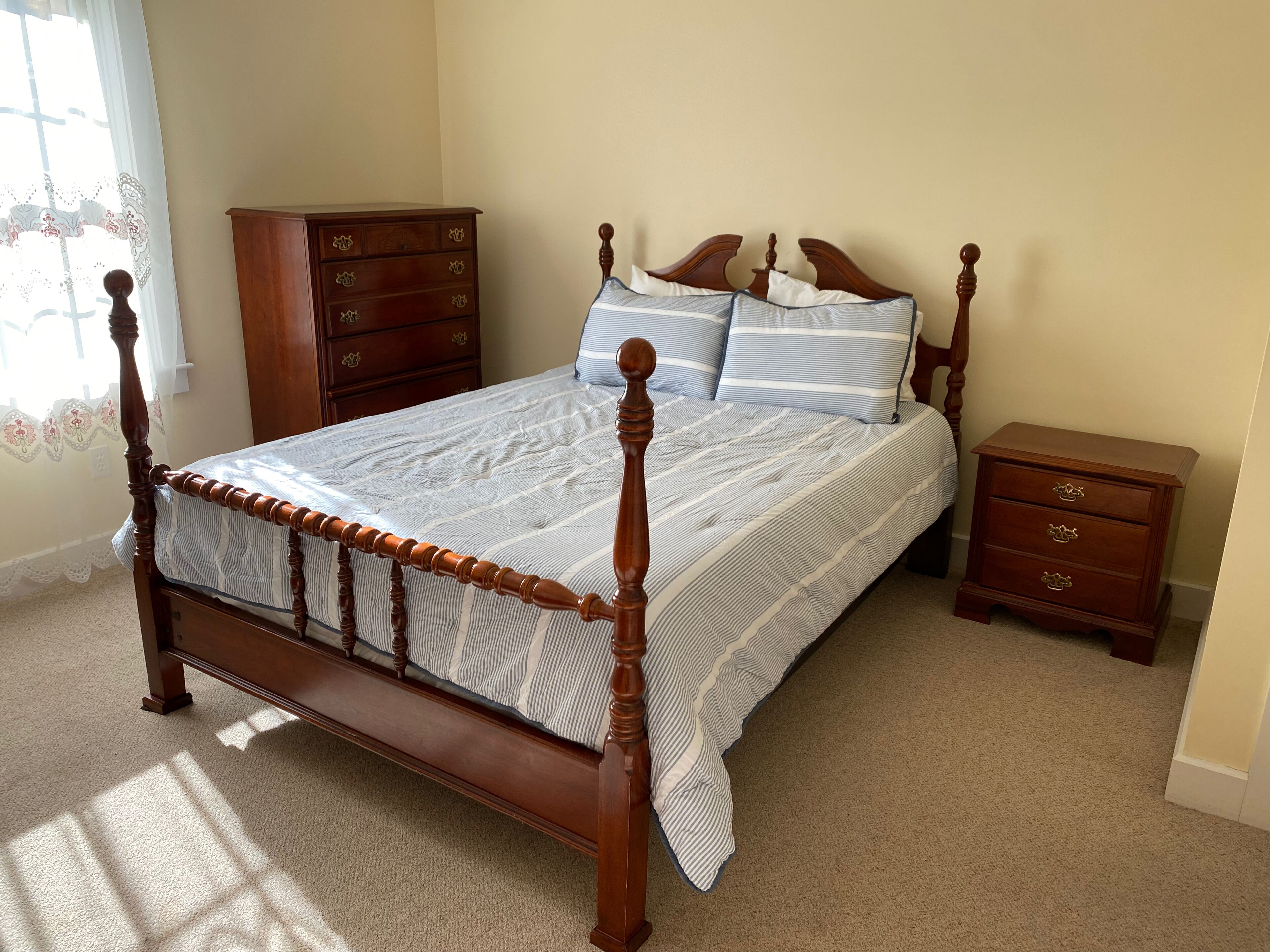 Quarto com cama de dossel de madeira, mesa de cabeceira, cômoda e janela com cortinas de renda.
