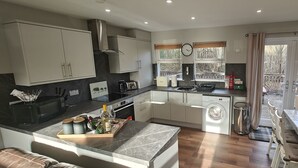 A modern kitchen with a built-in washing machine, a dining area with a table and chairs, and a view of the outside. - Ben Lomond Cottage - Loch Lomond and Arrochar Alps (Arrochar)