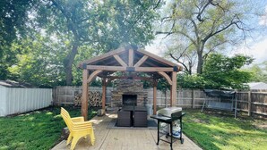 A backyard with a wooden pergola, a stone fireplace, a grill, and a yellow chair.