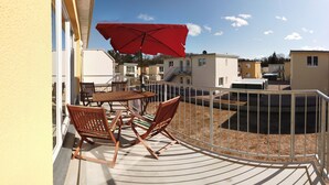 Een balkon met een tafel en stoelen, een rode parasol en uitzicht op omliggende gebouwen.