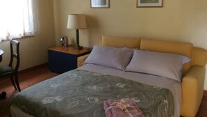 Une chambre d’hôtel avec un lit, une table de chevet avec une lampe, une chaise et deux tableaux encadrés au mur.