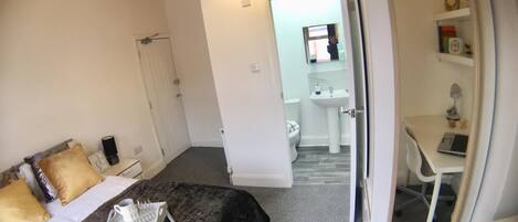 A bedroom with a bed, bedside table, and a mirror. There is a bathroom with a toilet, sink, and mirror. A desk with a chair is visible in the corner.