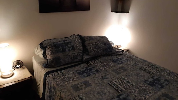 Un dormitorio con una cama, una mesita de noche con una lámpara, un florero en la pared y un cuadro con un atardecer sobre un puente.