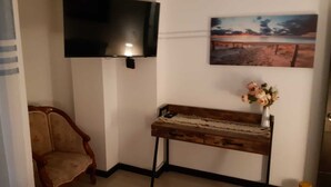 Una habitación con una televisión de pantalla plana montada en la pared, una mesita consola de madera con un jarrón de flores y una silla de estilo antiguo.