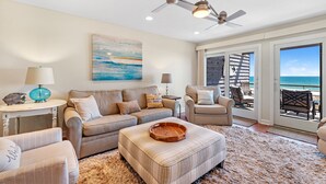 Ein Wohnzimmer mit Sofa, Hocker und Sesseln, einem Deckenventilator und Meerblick durch große Fenster.