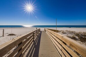 Une promenade en bois qui mène à une plage, avec un ciel dégagé et un soleil brillant.