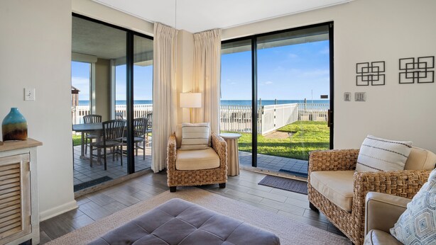 A spacious living area with a view of the sea, a wicker sofa, a cushioned armchair, and a dining table with chairs. - Romar Place 104 |  Beachfront Condo | Indoor & Outdoor Pools | Beach Getaways (Orange Beach)