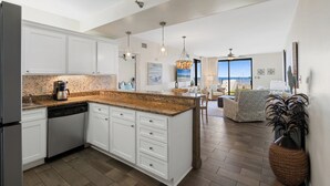 A modern kitchen with white cabinets, a granite countertop, and a dishwasher. There is a dining area with a view of the ocean. - Romar Place 104 |  Beachfront Condo | Indoor & Outdoor Pools | Beach Getaways (Orange Beach)