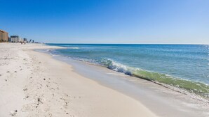 A beach with a clear sky, sandy shore, and buildings in the background. - Romar Place 104 |  Beachfront Condo | Indoor & Outdoor Pools | Beach Getaways (Orange Beach)